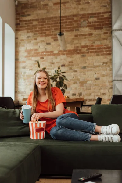 Allegra ragazza tenendo tazza usa e getta vicino popcorn e guardare film in soggiorno — Foto stock
