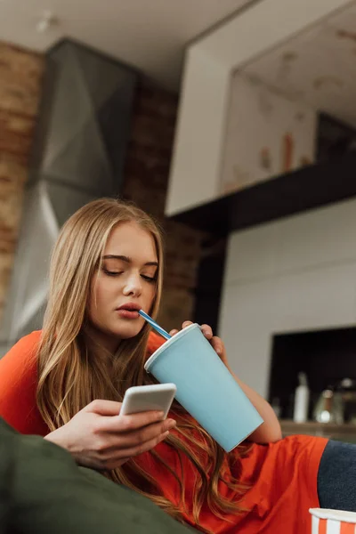 Giovane donna in possesso di tazza di carta con soda e utilizzando smartphone a casa — Foto stock