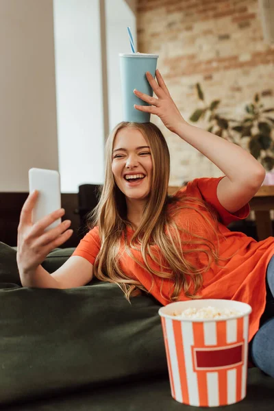 Messa a fuoco selettiva di allegra ragazza tenendo tazza di carta sopra la testa e prendendo selfie vicino secchio popcorn — Foto stock