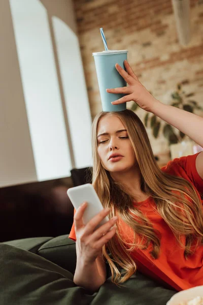 Foco seletivo de menina atraente segurando copo de papel acima da cabeça e tomando selfie perto de balde de pipoca — Fotografia de Stock