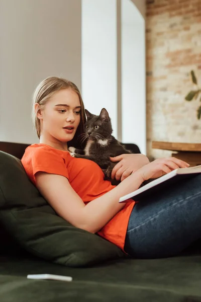 Foco seletivo de menina bonita segurando gato e livro de leitura na sala de estar — Fotografia de Stock