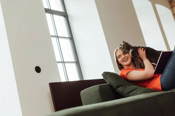 Enfoque selectivo de alegre joven mujer sosteniendo gato y mirando hacia otro lado en sala de estar - foto de stock