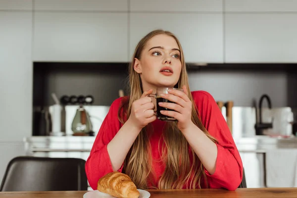 Jovem mulher segurando xícara de café perto de croissant saboroso na mesa — Fotografia de Stock