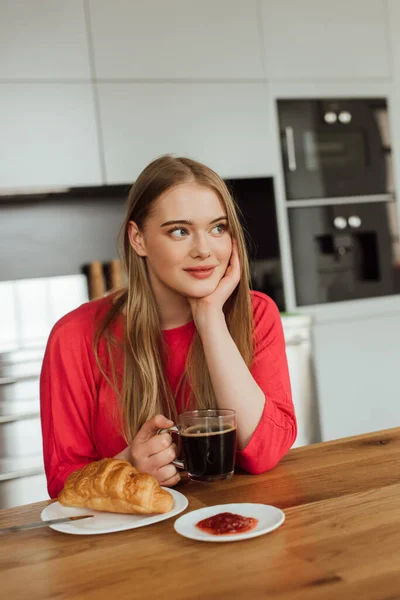 Menina sonhadora segurando xícara de café perto de croissant saboroso na mesa — Fotografia de Stock
