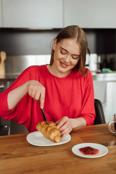 Happy girl holding knife and cutting tasty croissant on plate — Stock Photo