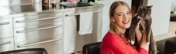 Concepto panorámico de mujer joven feliz sosteniendo gato lindo en la cocina - foto de stock