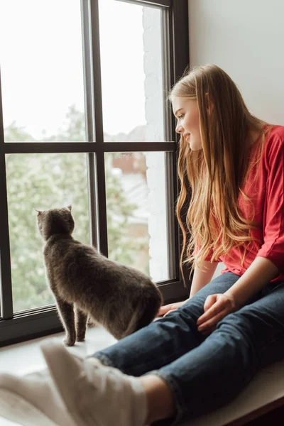 Foco seletivo da mulher feliz olhando para a janela perto de gato bonito — Fotografia de Stock
