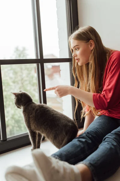 Selective focus of young woman pointing with finger at window near cute cat — Stock Photo