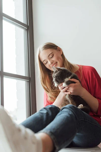 Enfoque selectivo de la mujer positiva sosteniendo lindo gato mientras está sentado en ventana alféizar - foto de stock