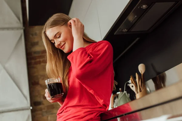 Enfoque selectivo de la mujer alegre sosteniendo taza de café y tocando el cabello - foto de stock
