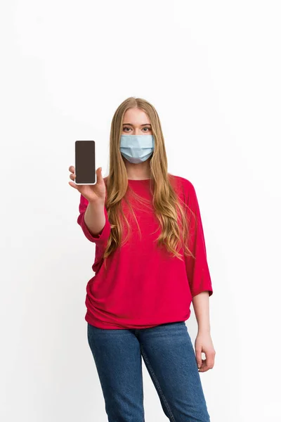 Mujer joven con máscara médica sosteniendo teléfono inteligente con pantalla en blanco aislado en blanco - foto de stock