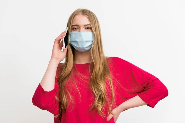 Giovane donna in maschera medica che parla su smartphone e in piedi con mano sull'anca isolata su bianco — Foto stock