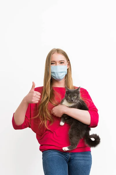 Молодая женщина в медицинской маске, держа в руках милую кошку и показывая большой палец, изолированный на белом — стоковое фото