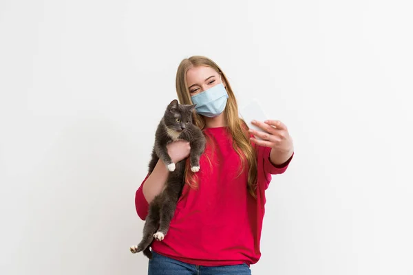 Jeune femme en masque médical tenant dans les bras chat mignon et prendre selfie isolé sur blanc — Photo de stock