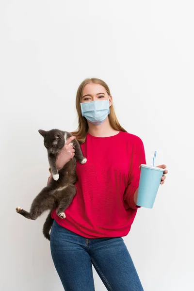 Young woman in medical mask holding in arms fluffy cat and disposable cup with soda isolated on white — Stock Photo