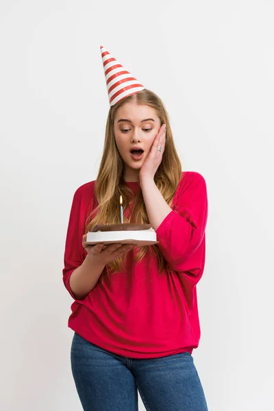 Chica sorprendida en gorra de fiesta mirando pastel de cumpleaños aislado en blanco - foto de stock