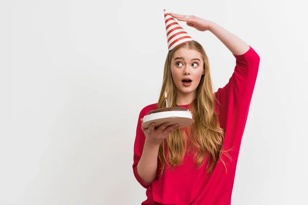 Surprised girl touching party cap and holding birthday cake isolated on white — Stock Photo