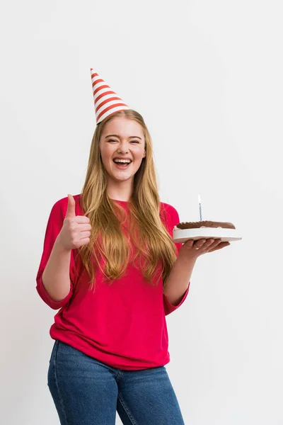 Alegre chica en partido gorra mostrando el pulgar hacia arriba y la celebración de pastel de cumpleaños aislado en blanco - foto de stock