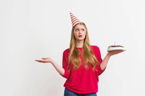 Confused woman in party cap holding birthday cake isolated on white — Stock Photo