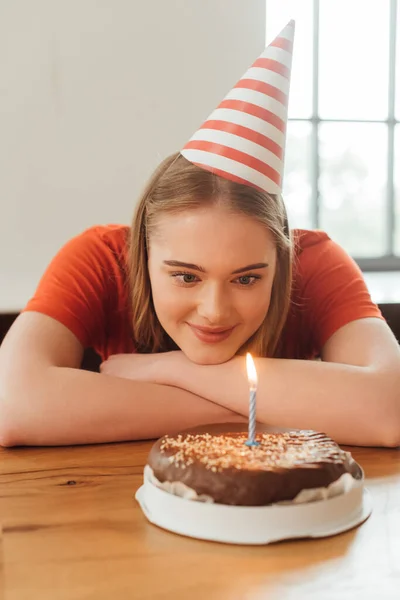 Selektiver Fokus der gut gelaunten Frau mit Partymütze auf brennende Kerze auf leckerem Geburtstagskuchen — Stockfoto