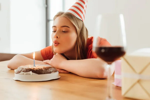 Selective focus of girl in party cap blowing out candle on birthday cake near presents and wine glass on table — Stock Photo