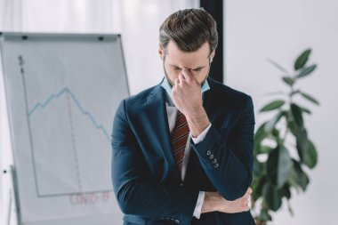 depressed businessman standing with bowed head and closed eyes near flipchart with graphs showing recession clipart