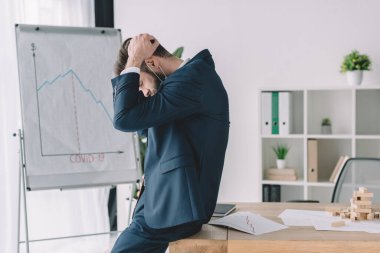 side view of depressed businessman holding hands on bowed head while near flipchart and workplace in office clipart