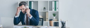 panoramic shot of upset businessman touching forehead while talking on smartphone at workplace clipart