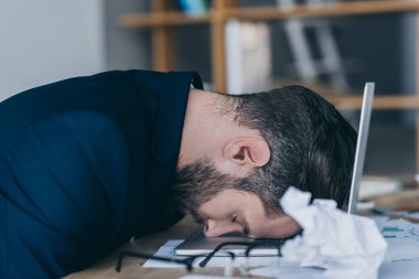 depressed businessman with closed eyes sitting at workplace with head on laptop near crumpled paper clipart