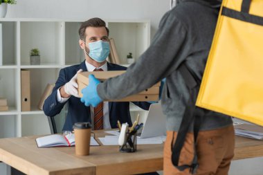 partial view of food delivery man giving pizza boxes to businessman in medical mask in office clipart