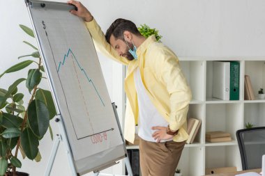 stressed businessman standing with closed eyes near flipchart with covid-19 inscription and graphs showing decrease clipart