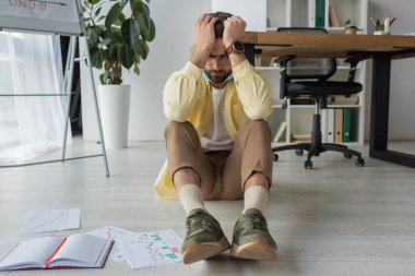 Depresyonda olan işadamı, elleri başının üstünde otururken yerde gözleri kapalı, çizelgeleri olan kağıtlar da defterin yanında oturuyor.