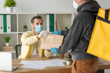 Tıbbi maske ve lateks eldiven takan iş adamına kağıt torba veren yemek dağıtım elemanı görüntüsü.