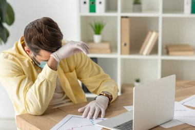 Medikal maskeli ve lateks eldivenli üzgün işadamı dizüstü bilgisayarın yanında oturuyor ve çizelgeli kağıtlar.