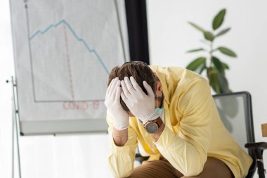 depressed businessman in latex holding hands on head while sitting near flipchart with covid-19 inscription infographics showing decrease clipart