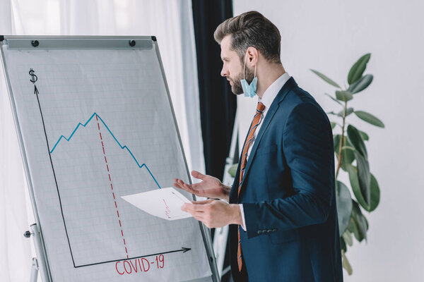 serious, thoughtful businessman holding paper while standing at flipchart with graphs showing decrease