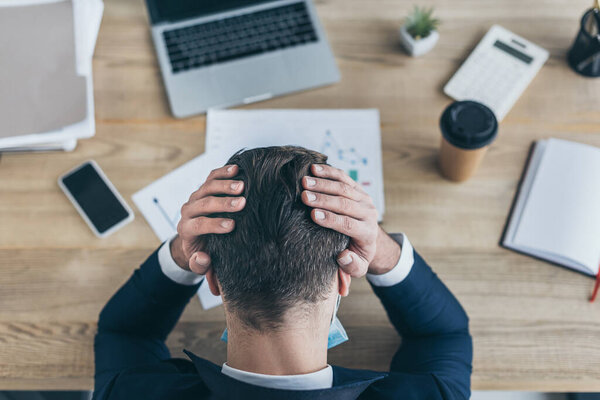 top view of depressed businessman covering head with hands while sitting at workplace near gadgets and documents