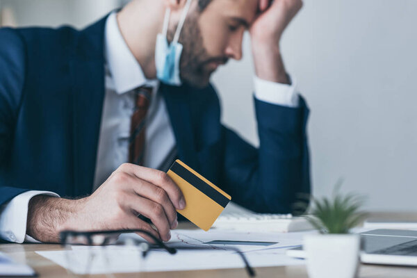 selective focus of upset businessman holding credit card while sitting at workplace with closed eyes and bowed head