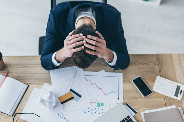 top view of depressed businessman holding hands on bowed head while sitting at desk with documents