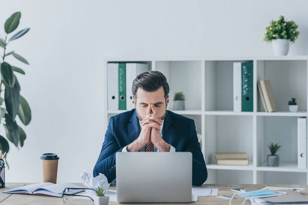depressed businessman holding hands near face while sitting at workplace with closed eyes