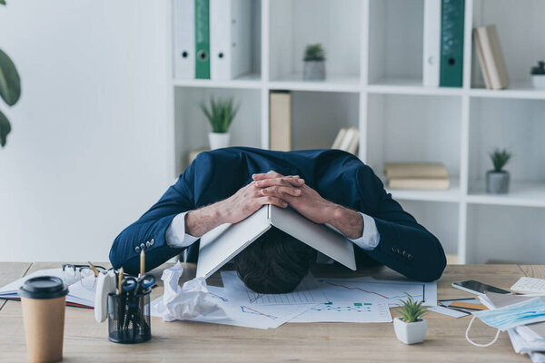 depressed businessman sitting at workplace and covering head with laptop