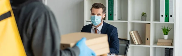 オフィスの職場に座っている医療マスクのビジネスマンの近くにピザボックスを保持する食品配達の男のクロップドビュー パノラマショット — ストック写真