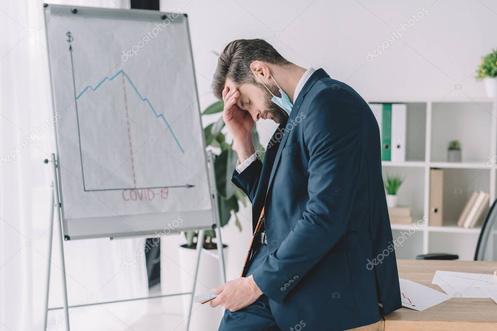 side view of stressed businessman standing with bowed head and closed eyes near flipchart with graphs showing recession