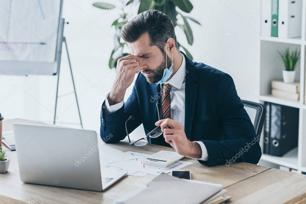 depressed, exhausted businessman holding eyeglasses and touching face while sitting at workplace near laptop and documents
