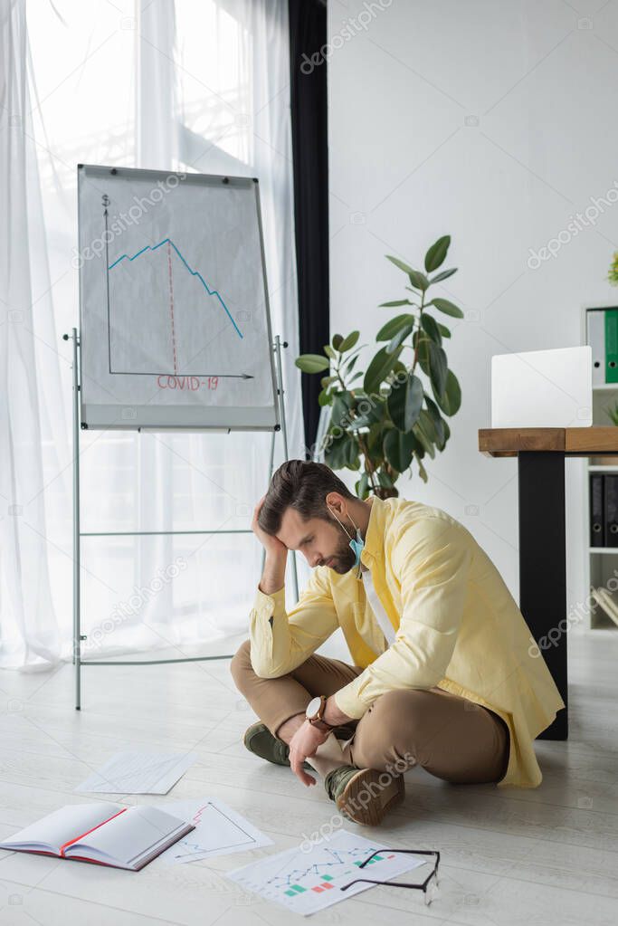 depressed businessman touching head while sitting on floor near documents and flipchart with covid-19 inscription and graphs showing recession 