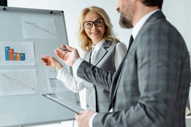 Elinde pano tutan işadamının yanındaki beyaz tahtadaki çizelgeleri gösteren gülümseyen iş kadınının seçici odak noktası 