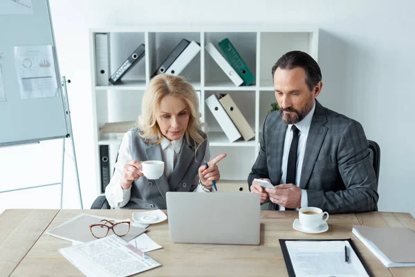 Bizneswoman Filiżanką Kawy Wskazując Palcem Biznesmena Smartfonem Pobliżu Laptopa Dokumentów — Zdjęcie stockowe