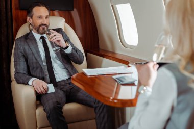 Selective focus of smiling businessman with champagne looking at businesswoman in airplane   clipart
