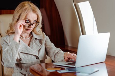 Selective focus of businesswoman holding eyeglasses while using laptop near credit cards and passports with air tickets on table in airplane  clipart