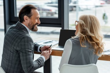 Gülümseyen işadamı, ofisteki evrak ve aletlerin yanındaki iş kadınına bakıyor. 
